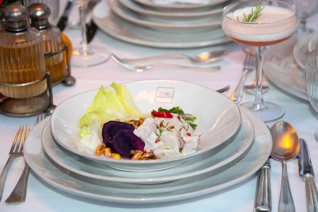 Plato de ceviche servido con lechuga, rodajas de batata morada y mazorcas de maíz, acompañado de un cóctel en una copa.