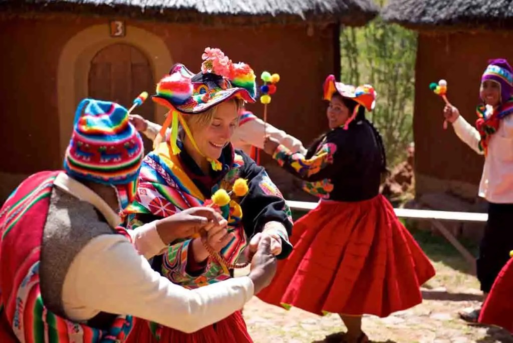 Grupo de personas bailando con trajes tradicionales en un entorno rural andino, con casas de adobe y techos de paja de fondo.