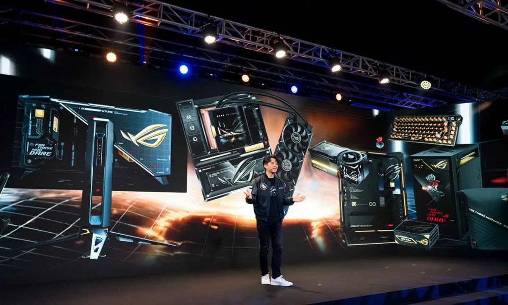 Presentador de tecnología en un evento, con una pantalla que muestra varios productos de hardware y gaming de la marca ROG.