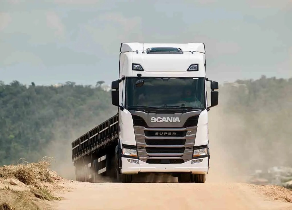 Camión Scania Super circulando por un camino de tierra, con polvo en el aire y un paisaje verde de fondo.