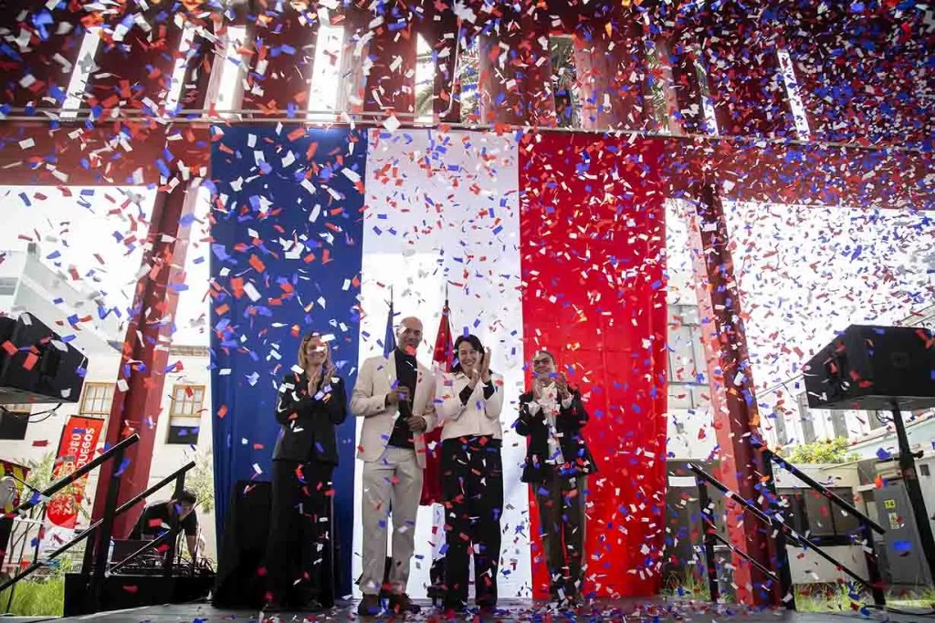 Celebración con confeti rojo, blanco y azul en un evento, con tres personas en el escenario aplaudiendo frente a un fondo decorado con los colores de la bandera.