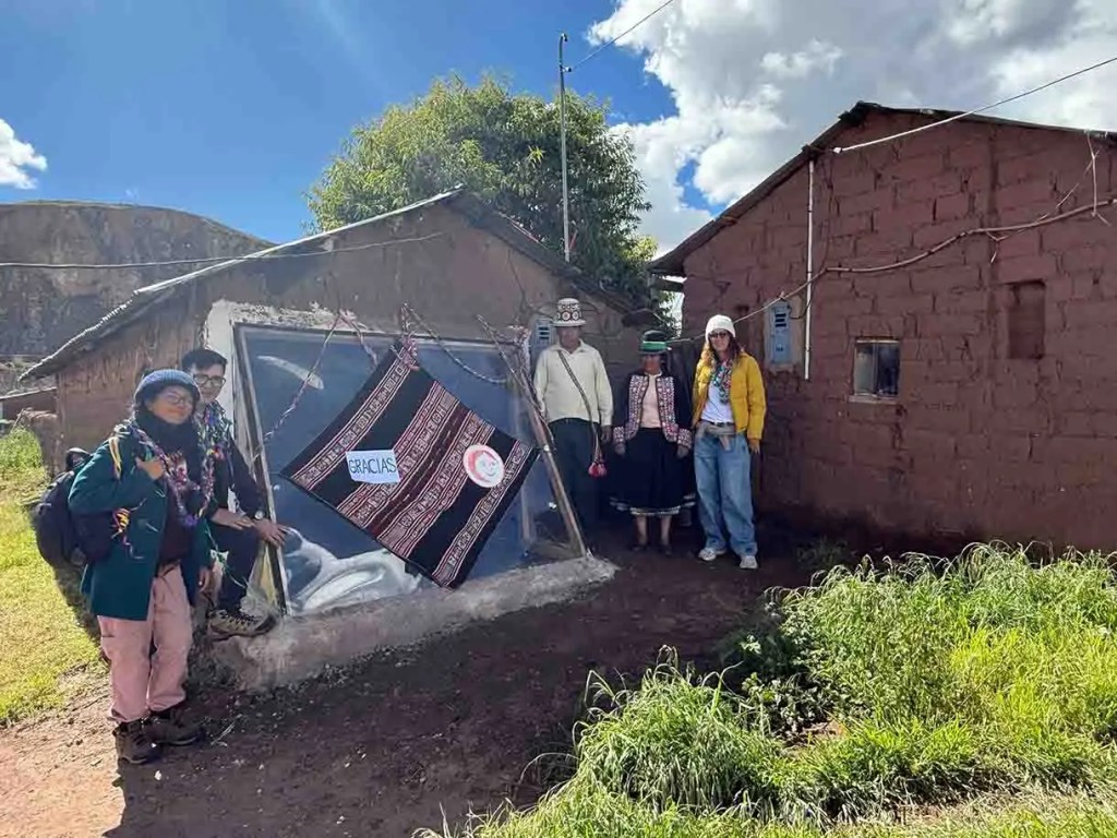 Grupo de personas posando frente a una casa de adobe con una ventana reflejante. Se observa un cartel que dice 'GRACIAS'.