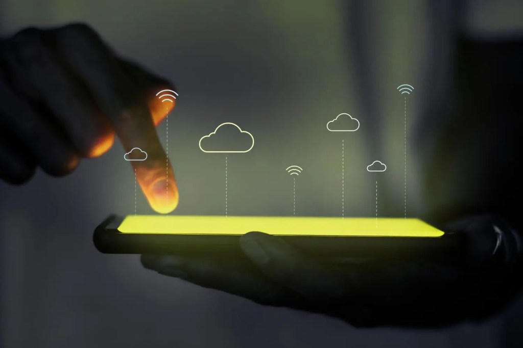 Mano tocando una pantalla de teléfono móvil amarilla, con iconos de nubes y conexión en la parte superior.