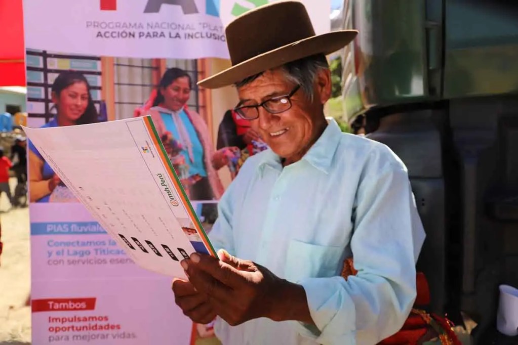 Hombre de edad avanzada con un sombrero, leyendo un documento en un evento comunitario, con un fondo que muestra información sobre un programa de inclusión social.