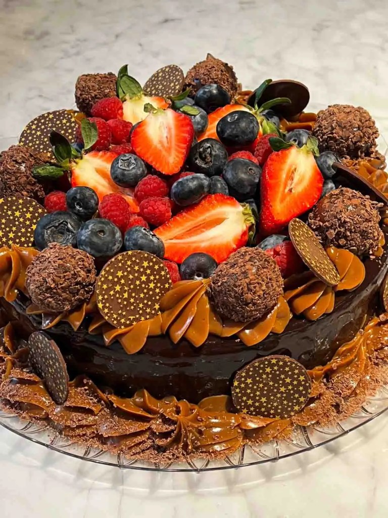Torta decorada con fresas, arándanos, frambuesas y trufas de chocolate, con detalles de chocolate y crema en la base.